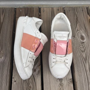 Valentino Garavani open sneakers  - Snygga Valentino Garavani open sneakers i vitt skinn med rosa och aprikosfärgade detaljer. Klassiska nitar baktill och breda band över snörningen ger en unik look. Perfekta för dig som vill sticka ut med exklusiva sneakers.