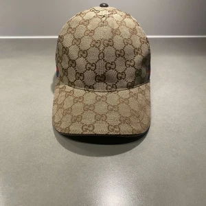Gucci keps med GG-mönster - Snygg keps från Gucci i beige och brunt med klassiskt GG-monogram över hela kepsen. Kepsen har justerbar rem bak i skinn och ikoniska gröna och röda ränder på sidan. Perfekt accessoar för dig som vill sticka ut med lyxig streetstyle.