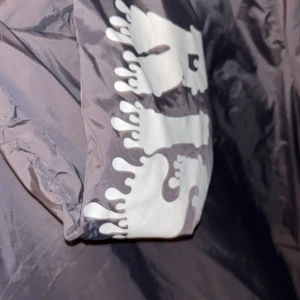 Svart vindjacka från risky  - Svart vindjacka från risky med vit tryckt logga på ärmen. Jackan har huva med dragsko, hel dragkedja framtill och elastiska muddar. Lätt och smidig modell i syntetmaterial, perfekt för blåsiga dagar. denna varan säljs ej längre