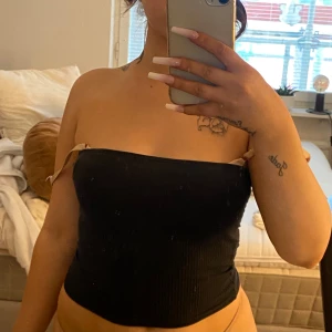 Svart ribbad bandeau tubtopp - Säljer en svart ribbad bandeau tubtopp med knytband på sidorna. Toppen är tight och ärmlös, perfekt för sommardagar eller när du vill ha en enkel och stilren look. Passar dig som gillar minimalistisk stil och vill visa axlarna.