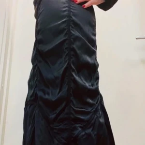 Svart långklänning By Malene Birger - Supersnygg svart långklänning från By Malene Birger i glansigt material. Klänningen har långa ärmar, markerad midja och coola rynkade detaljer längs hela längden. Perfekt för dig som vill sticka ut med en elegant och trendig look. Aldrig använd.