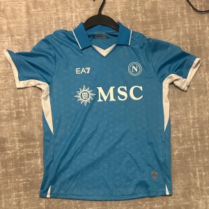 Napoli EA7 blå fotbollströja - Snygg Napoli fotbollströja från EA7 i blått med vita detaljer och tryck. Tröjan har korta ärmar, klassisk krage och är tillverkad i ett lätt, ventilerande material. MSC-logga och Napoli-emblem på bröstet, samt diskret mönster med små N över hela tyget.