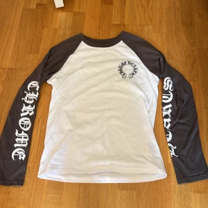 Chrome Hearts långärmad tröja - Säljer en långärmad tröja från Chrome Hearts med vita och mörkgrå kontrastärmar. Stora tryck med Chrome Hearts-logga på ryggen och ärmarna. Skön passform och tillverkad i bomull, perfekt för dig som gillar streetwear vibes.