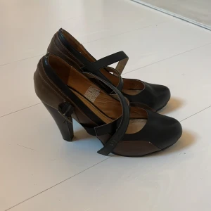Svarta och bruna klackskor från Tiamo - Snygga pumps från Tiamo i svart och brunt skinn med rund tå och hög klack. Skorna har två korsade remmar med spänne över foten för extra stilpoäng. Perfekta för dig som vill sticka ut med en klassisk men cool look.