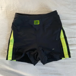 Svarta Adidas träningsshorts med neonränder - Svarta träningsshorts från Adidas med neon-gula ränder längs sidorna och Adidas-logga framtill. Tight passform och elastisk midja, perfekta för sport eller gym. Materialet är syntetiskt och snabbtorkande, vilket gör dem grymma för intensiva pass.
