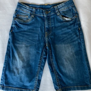 Blå jeansshorts från Xmax stl 128 - Snygga blå jeansshorts från Xmax med klassisk femficksdesign, gul kontrastsöm och knappstängning. Shortsen har normal passform och är perfekta för varma dagar. Materialet är slitstarkt denim och färgen har en cool tvättad look.