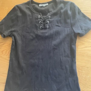 Svart snörad topp från KappAhl - Svart ribbad topp från KappAhl med korta ärmar och snygg snörning framtill vid halsen. T-shirten har en rundad halsringning och är gjord i mjuk bomull, perfekt för en chill och stilren look.