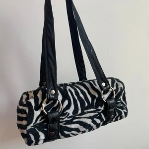 Zebramönstrad axelväska med ringdetaljer - Cool axelväska i svartvitt zebramönster med mjuk pälsliknande yta. Perfekt för dig som vill sticka ut med en trendig accessoar. Handtagen är lite slitna vilket man kan se på bilderna.