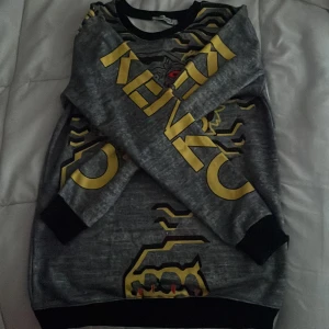 Grå Kenzo sweatshirt med tigertryck - Kenzo sweatshirt i grått med stort tigertryck och röda ögon framtill. Gula och svarta grafiska detaljer samt Kenzo-logga på bröstet och ärmarna. Långärmad modell med svarta muddar vid ärmslut och nederkant. Perfekt statement piece för dig som gillar streetstyle. Passar xs,aldrig andvänt nypris 1000