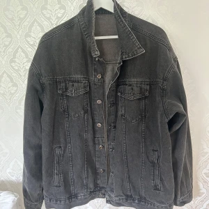 Svart oversized jeansjacka - Snygg svart jeansjacka i oversized modell med klassiska bröstfickor och knappar framtill. Jackan har långa ärmar och raka sömmar som ger en cool och avslappnad vibe. Perfekt att slänga över en hoodie eller t-shirt för en streetig look. Ej säker om storlek men den är oversized och passar M-L.