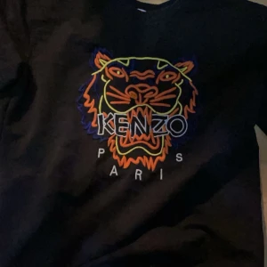 Svart Kenzo sweatshirt med tigertryck - Svart sweatshirt från Kenzo med ikoniskt broderat tigerhuvud i neonfärger och texten 'KENZO PARIS' framtill. Tröjan har rund halsringning och är långärmad. Perfekt statement-piece för dig som gillar streetstyle.