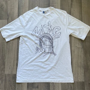 H&M vit NYC t-shirt - Säljer vit t-shirt med oversized passform från H&M DIVIDED. Det finns två små hål på framsidan av t-shirten. Modellen är 167 cm lång, väger 63 kg. T-shirten har längden 78 cm med axeln 49 cm.