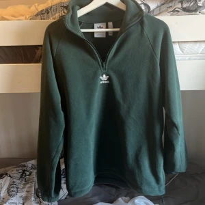 Grön fleece half zip från Adidas - Mörkgrön fleece-tröja från Adidas med broderad logga på bröstet och stor Adidas-tryck på ryggen. Tröjan har half zip, hög krage och långa ärmar. Perfekt för kyliga dagar och riktigt skön att ha på sig.