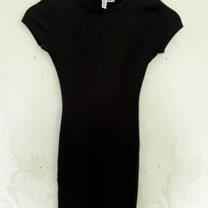 Svart ribbad kortklänning från Divided - En svart, tight ribbad kortklänning från Divided. Klänningen har korta ärmar och rund halsringning. Enkel och stilren modell som sitter figurnära.