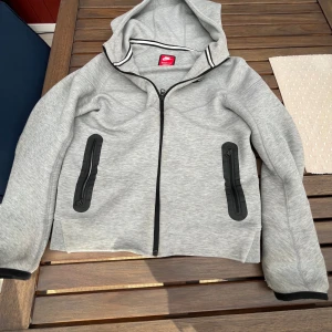 Grå hoodiejacka från Nike - Snygg grå hoodiejacka från Nike med hel dragkedja och svarta detaljer vid fickorna. Jackan har huva, långa ärmar och sportig look. Perfekt för chill dagar eller när du vill ha en enkel och stilren jacka. Materialet är mjukt och bekvämt.
