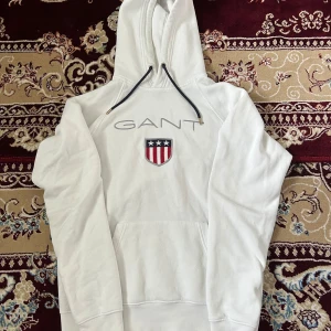 Vit hoodie från GANT med logga/ även byte - Vit hoodie från GANT med broderad logga och sköld på bröstet. Tröjan har huva med svarta snören och en stor magficka. Klassisk och stilren design, perfekt för dig som gillar preppy streetstyle.