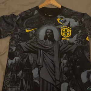 Brasil Nike fotbollströja svart/gul - Snygg Brasilien fotbollströja från Nike med unikt svart och grått grafiskt tryck av Kristusstatyn och brasilianska motiv. Gula detaljer, CBF-emblem på bröst och rygg, samt Nike-logga. Tillverkad i lätt och ventilerande material, perfekt för match eller träning.