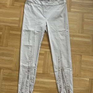 Beige byxor med volangdetaljer Gustav - Snygga beige byxor från Gustav med hög midja och smal passform. Byxorna har unika volangdetaljer och knappar längs benen framtill, vilket ger en cool och annorlunda look. Tillverkade i ett mjukt bomullsmaterial som känns skönt mot huden. Från danska Gustav 