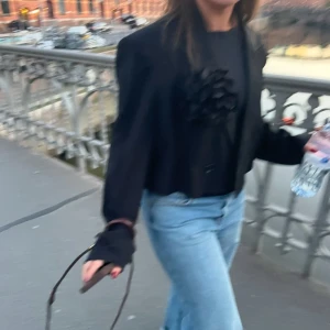 Svart croppad kavaj - Säljer en svart croppad kavaj med lång ärm.  Jackan har klassisk krage, knäppning framtill och en stilren, loose passform. Perfekt för dig som vill ha en trendig och unik look. (Inte blomman) köpt i större storlek så man skulle få en over sized look