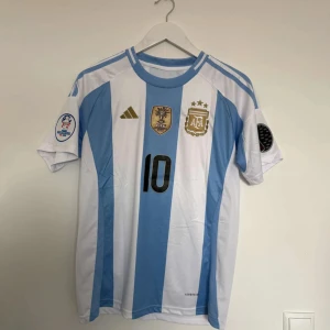 Argentina Messi 10 Copa America 2024 tröja - Säljer en officiell Argentina fotbollströja från Adidas med Messi 10 på ryggen. Tröjan har klassiska blå och vita ränder, broderade guldiga mästerskapsmärken och Copa America 2024-patch på ärmen. Tillverkad i lätt och ventilerande polyester, perfekt för match eller support.            Prislappen sitter kvar!!!