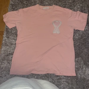 Rosa t-shirt från Ami med hjärtdetalj - Snygg rosa t-shirt från Ami Alexandre Mattiussi med korta ärmar och ett stort broderat hjärta med bokstaven A på bröstet. Tillverkad i mjuk bomull och har en klassisk passform. Perfekt för dig som gillar stilrena plagg med en twist.