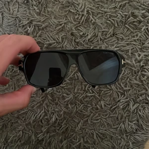  Tom Ford solglasögon  - Snygga svarta solglasögon från Tom Ford med klassisk form och mörka glas. Ramen är i blank svart plast med guldiga T-detaljer på sidorna. Perfekt accessoar för en cool look och tillverkade i Italien. Kan gå ner i pris vid snabb affär