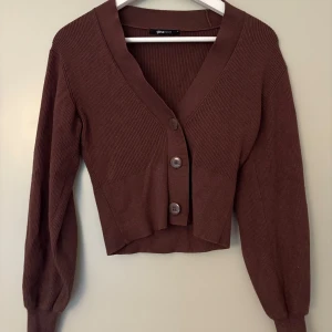 Brun croppad kofta från Gina Tricot - Säljer en brun croppad kofta från Gina Tricot med v-ringning och stora knappar framtill. Koftan är ribbstickad och har långa ärmar med breda muddar. Perfekt att slänga över en topp för en chill look. Använd fåtal gånger.