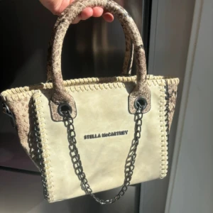  handväska Stella McCartney - Snygg beige handväska från Stella McCartney med detaljer i ormskinnsmönstrad syntet och svarta kedjehandtag. I gott skick men finns tecken på användning ☺️
