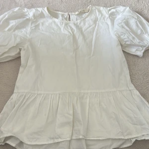 Vit blus med volang och puffärm - Supersöt vit blus med rund halsringning, puffiga korta ärmar och volang nertill. Blusen har en luftig och lätt känsla, perfekt för en fräsch och stilren look. Passar till både jeans och kjol för en trendig outfit.