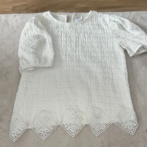 Vit blus med spetsdetaljer från Zara - Superfin vit blus från Zara med korta puffärmar och unika spetsdetaljer på både ärmar och nederkant. Blusen har en vågig struktur i tyget och spetsen har ett snyggt geometriskt mönster. Perfekt för dig som gillar uppklädda och trendiga plagg.