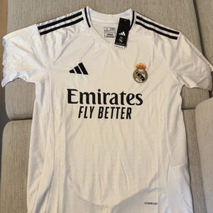Real Madrid hemmatröja Adidas vit - Snygg Real Madrid hemmatröja från Adidas i vitt med svarta detaljer och klubbmärke på bröstet. Tröjan har korta ärmar, klassiska svarta ränder på axlarna och är tillverkad i lätt, ventilerande material med Aeroready-teknologi. Perfekt för fotbollsälskare!