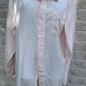 Ljusrosa skjorta med bröstficka - En stilren ljusrosa skjorta som endast användes 1 gång. Skjortan har en bröstficka och långa ärmar, perfekt för en clean och avslappnad look.                                                       Passar dig som gillar enkel och tidlös stil.                                                        En liten gul fläck finnes men endast på insidan av skortan så den syns inte 