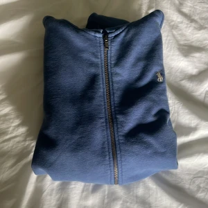 Blå hoodie från Polo Ralph Lauren - Snygg blå hoodie från Polo Ralph Lauren med dragkedja och huva. Klassisk logga broderad på bröstet och praktiska fickor framtill. Tillverkad i mjuk bomull, perfekt för chill dagar. Stilsäker och enkel att matcha med jeans eller joggers. Kan pruta lite på priset. Äkta👍