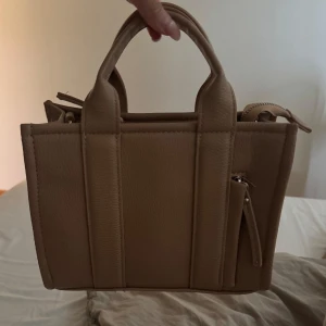 Beige handväska i skinnlook - Stilren beige handväska med två  handtag  Väskan har en boxig form och är tillverkad i material som liknar skinn. Perfekt storlek för att bära med sig det viktigaste och passar till många outfits.