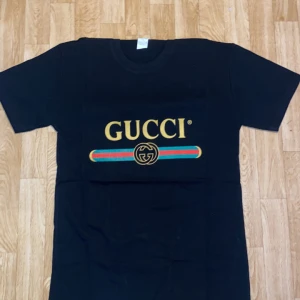 Gucci t shirt S/M - Svart t-shirt från Gucci med ikonisk guldig logga framtill och färgglada ränder i grönt och rött. Klassisk passform med rund hals och korta ärmar. Perfekt statement-plagg för dig som vill sticka ut med lyxig streetstyle.