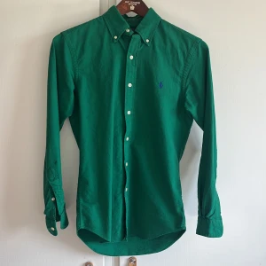 Grön skjorta från Polo Ralph Lauren XS - Snygg grön skjorta från Polo Ralph Lauren i custom fit. Klassisk button-down krage, vita knappar och lång ärm. Broderad blå logga på bröstet. Skjortan är i bomull och passar perfekt till jeans eller chinos. Endast en liten defekt på ena armen där det är en svart fläck