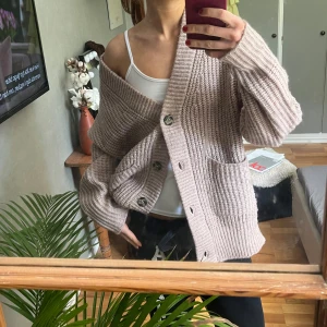 Beige oversized stickad kofta - Mysig oversized kofta i beige stickat material med stora knappar framtill och två rejäla fickor. Koftan har lång ärm och bred ribbad kant längs öppningen. Perfekt att slänga på sig över ett linne för en chill look.