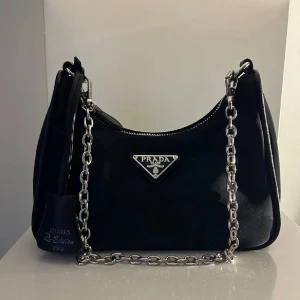 Pradaväska  - Snygg svart Prada väska. Väskan har det klassiska Prada-emblemet framtill och en liten avtagbar miniväska på remmen. Tillverkad i nylon med dragkedja upptill. 