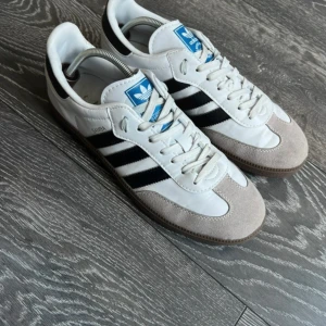 Adidas Samba - Klassiska Adidas Samba sneakers i vitt skinn med svarta ränder och detaljer, grå mocka vid tån och brun gummisula. Ikonisk blå Adidas-logga på plösen och guldfärgad Samba-text på sidan. Perfekta för en sportig och stilren look.