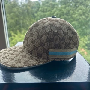 Gucci keps  - Snygg keps från Gucci i beige och brunt med klassiskt GG-mönster över hela kepsen. Har en ljusblå och beige rand på sidan samt ljusblå justerbar rem bak. Perfekt accessoar för att lyfta din outfit.