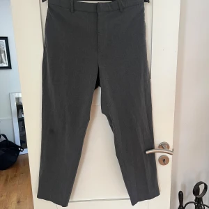 Grå kostymbyxor från Uniqlo M - Snygga grå kostymbyxor från Uniqlo i storlek M. Byxorna har klassisk rak passform, bälteshällor och stängs med dragkedja och knapp. Materialet är en mix av polyester, viskos och elastan vilket gör dem både bekväma och stilrena.