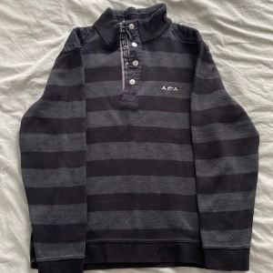 Randig pikétröja Mise au Green M - Snygg randig pikétröja från Mise au Green i svart och grått. Tröjan har hög krage, knappar framtill och diskret logga på bröstet. Tillverkad i mjuk manchester med långa ärmar och ribbade detaljer vid kragen. Perfekt för dig som gillar en chill men stilren look.