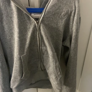 Grå Juicy Couture hoodie med dragkedja - Grå hoodie från Juicy Couture med broderad JC-logga på bröstet. Tröjan har huva, fickor framtill och hel dragkedja med snygg J-detalj. Perfekt för chill dagar och streetstyle. Mjuk och skön i bomullsmix, långärmad och klassisk passform.