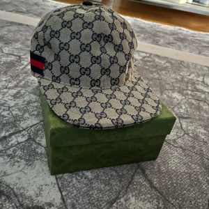 Gucci keps med GG-mönster och röd detalj - Snygg keps från Gucci med klassiskt GG-monogram i mörkblått på beige botten. Kepsen har en röd och marinblå rand på sidan och justerbar rem baktill. Tillverkad i bomullscanvas med mörkblått innerfoder. Perfekt för dig som vill sticka ut med lyxig streetstyle.