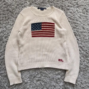 Beige stickad tröja Ralph Lauren Sport - Beige stickad tröja från Ralph Lauren Sport med amerikansk flagga broderad på bröstet och RL-broderi nedtill. Klassisk rund halsringning och ribbade muddar. Perfekt för dig som gillar preppy och tidlös stil.