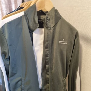 Grön vindjacka från Hang With Me + byxor - Sportig grön vindjacka från Hang With Me med vita detaljer längs ärmarna och trycket 'Urban Future Young Leaders' på bröstet. Jackan har dragkedja framtill, ståkrage och praktiska fickor. Perfekt för dig som gillar en clean och modern stil.