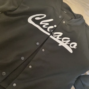 Svart bomberjacka med Chicago-tryck - Snygg svart bomberjacka med vit text 'Chicago' över bröstet. Jackan har knappar framtill och klassisk ribbad krage och muddar. Perfekt för dig som gillar sportig streetstyle och vill sticka ut med en enkel men cool look.