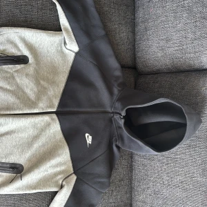 Nike hoodiejacka svart/grå med dragkedja - Nike hoodiejacka i svart och ljusgrått med hel dragkedja framtill och två fickor med dragkedja. Jackan har huva och sportig look, perfekt för chill eller träning. Snygg kontrast mellan färgerna och klassisk Nike-logga på bröstet.