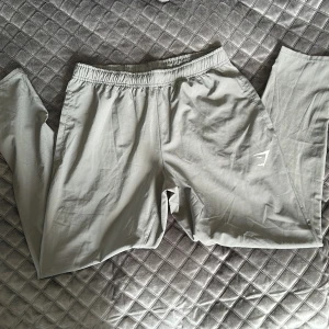 Grå träningsbyxor från Gymshark S - Snygga grå träningsbyxor från Gymshark i storlek S. Byxorna har elastisk midja och är tillverkade i ett mjukt och lätt syntetmaterial. Perfekta för gymmet eller chill hemma. Diskret Gymshark-logga på benet för en clean look.