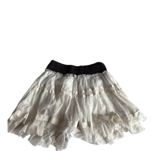 Vita volangskjorts med bred midjeresår - Supersöt vit kortkjol med flera lager volanger och bred svart resår i midjan. Kjolen har spetsdetaljer och ett skirt, lätt material som ger en fluffig känsla. Perfekt för dig som gillar romantisk och lekfull stil.